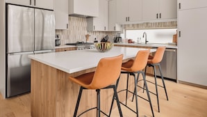 Eine moderne Küche mit weißen Schränken, einem Edelstahlkühlschrank, einer Holzinsel mit Barhocker und einer Pendelleuchte.