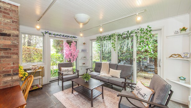 A cozy living room with a sofa, chairs, a coffee table, and a brick wall. There are large windows with plants and flowers. - Spacious 5-Bedroom 6 Bed House In Gorgeous Upper Montclair with AC, Theater Room (Upper Montclair)