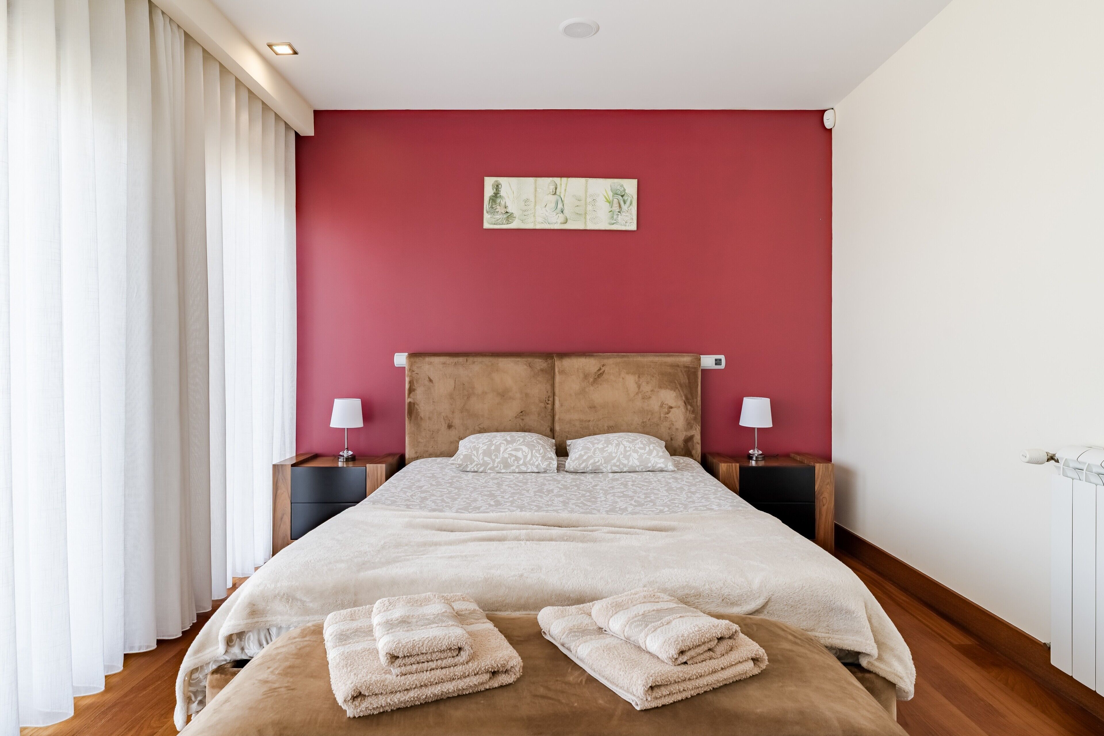 Een slaapkamer met een groot bed, twee nachtkastjes met lampen, een rode wand en een raam met witte gordijnen.