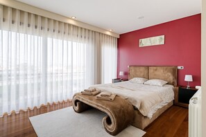 Un dormitorio con una cama grande, un cabecero de madera, una mesita de noche con lámpara y vistas a través de cortinas translúcidas.