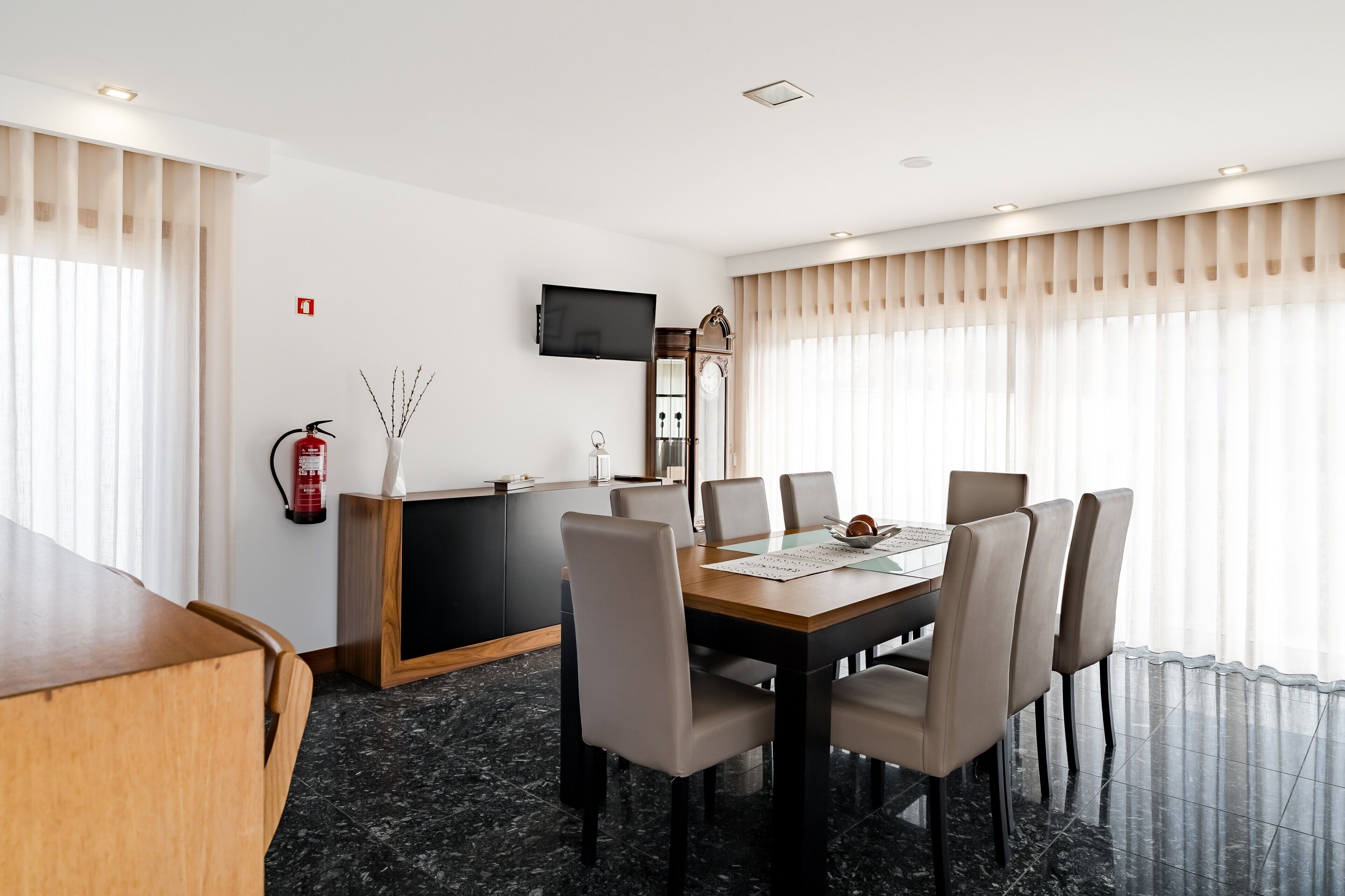 Un comedor de hotel con una mesa de madera, seis sillas, un televisor montado en la pared y un extintor en la pared.