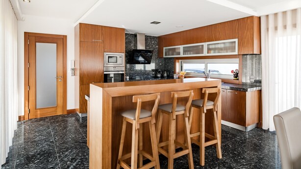 A modern kitchen with wooden cabinets, a marble countertop, a built-in oven, and a bar area with stools. - Casa Do Chino - Suite with Pool View (Belinho)