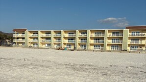 A multi-story building with balconies, a sandy ground, and a clear sky.