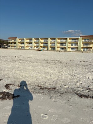 A multi-story building with balconies, a sandy ground, and a clear sky.