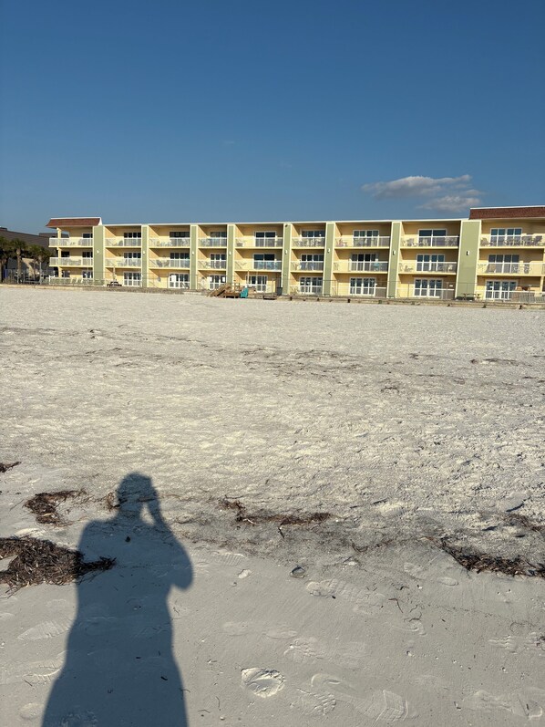 A multi-story building with balconies, a sandy ground, and a clear sky.