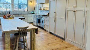 Een keuken met een groot eiland, houten krukken, witte kastjes en uitzicht op buiten.