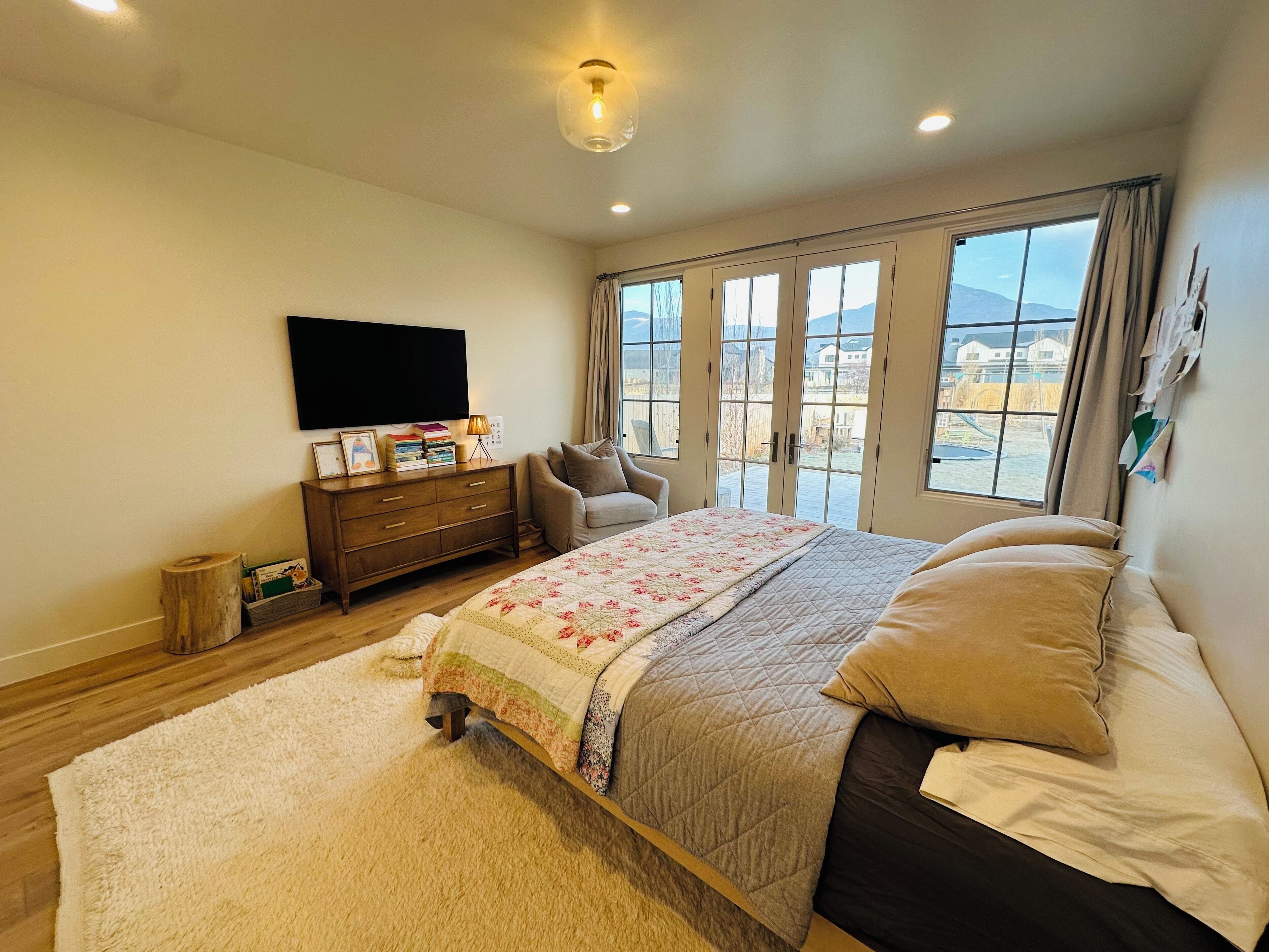 Un dormitorio con cama, cómoda, televisor y ventanas grandes.