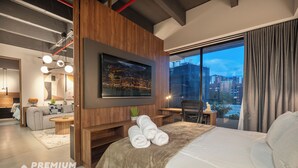Een moderne hotelkamer met een groot bed, een flatscreen televisie aan de muur, een zithoek met een bank en een kleintje tafeltje, en een uitzicht op de stad door grote ramen.