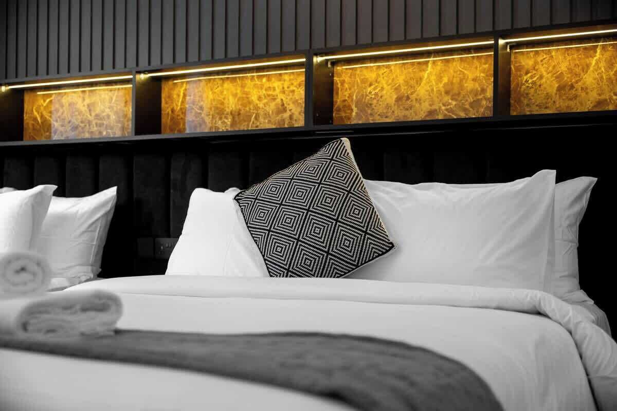 A hotel room with a modern design, featuring a large bed with white bedding and a geometric-patterned pillow, a dark headboard with a marble-like texture, and a wall with illuminated panels.