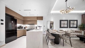 A modern kitchen with a central island, a dining area with a round table, and a living space with a sofa and a chandelier. - Cozy 3BR nr Circle Mall Sleeps 12 | Pool & Sauna (Dubai)