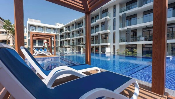Zona de piscina de hotel con múltiples sillas de descanso y un edificio con balcones al fondo.