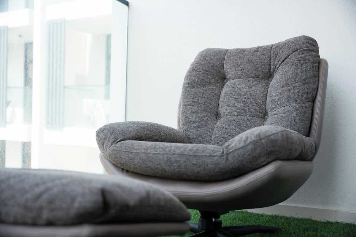 Un fauteuil moderne doté d’un haut dossier et d’un siège rembourré, placé dans une pièce avec une grande fenêtre et un sol recouvert d’un gazon artificiel vert.