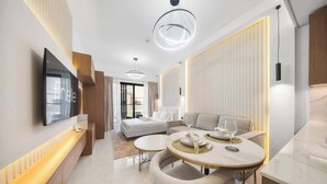 Una habitación de hotel moderna con televisión de pantalla plana, un comedor con una mesa redonda y sillas, un sofá y un ventanal grande.