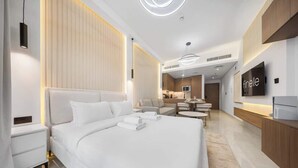 Habitación de hotel moderna con una cama grande, mesitas de noche, televisión y una kitchenette al fondo.