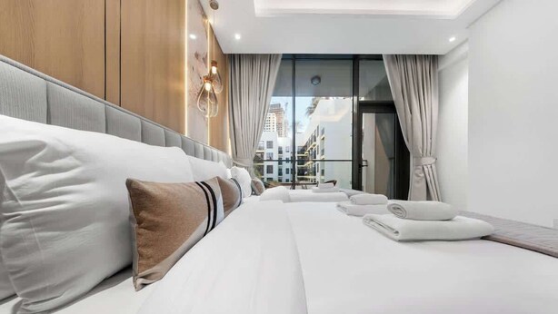 A modern hotel room with a large bed, a view of the city through floor-to-ceiling windows, and a balcony with a cityscape view. - JVC 1BR nr Circle Mall | Sleeps 7 | Pool & Gym (Dubai)