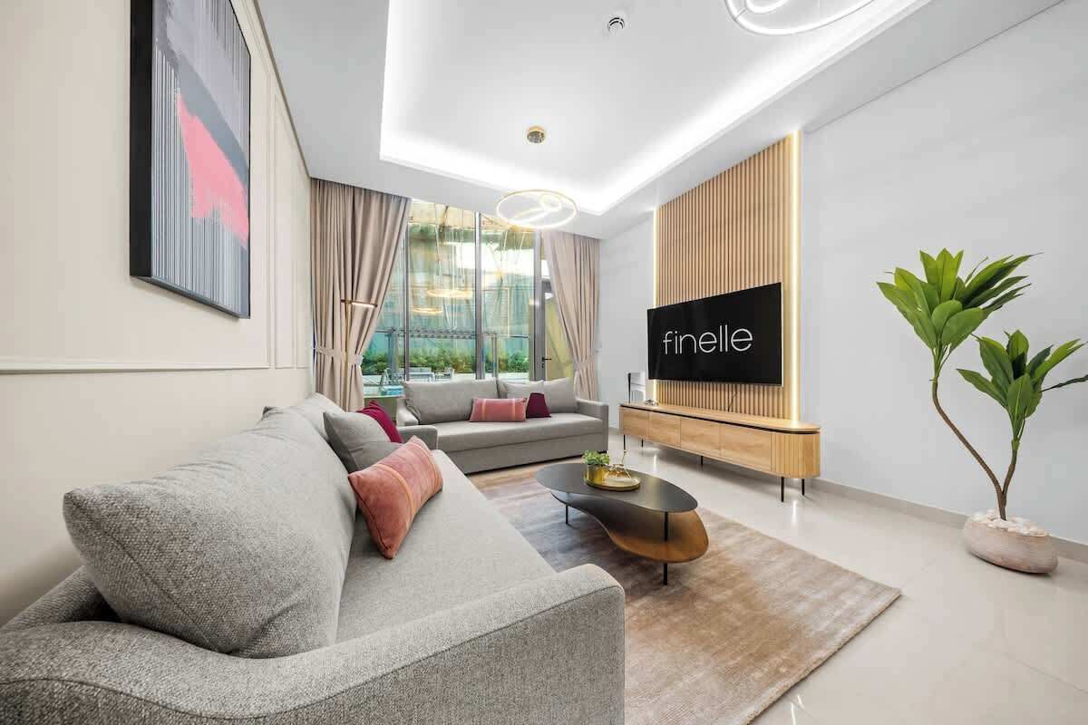 A modern living room with a grey sofa, a coffee table, a TV on a wooden stand, and a large window with curtains.