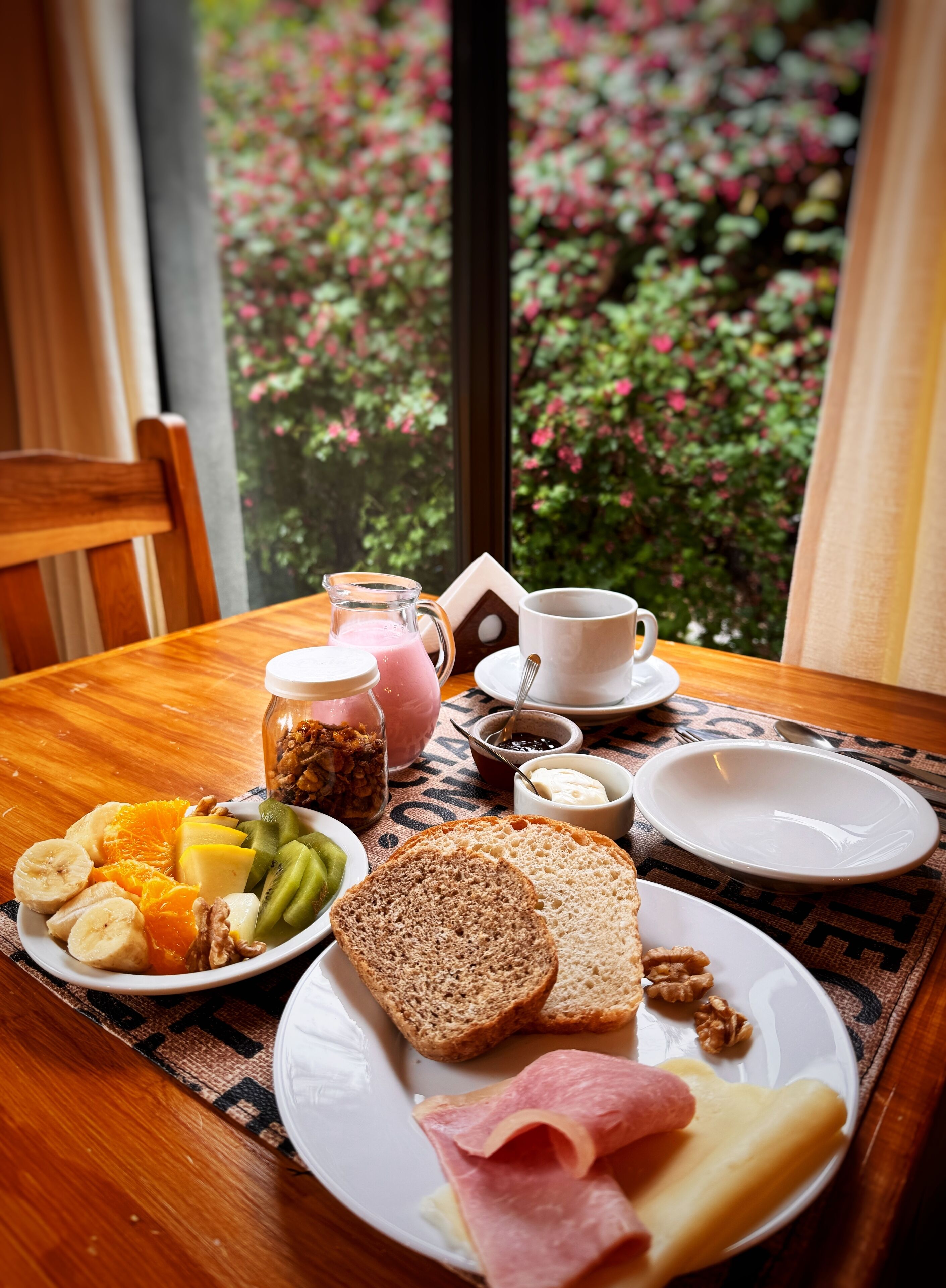 Une table de petit-déjeuner dressée avec une variété d’aliments, notamment du pain, du fromage, du jambon et des fruits, le tout mis en valeur par un décor composé d’une fenêtre donnant sur des fleurs roses.