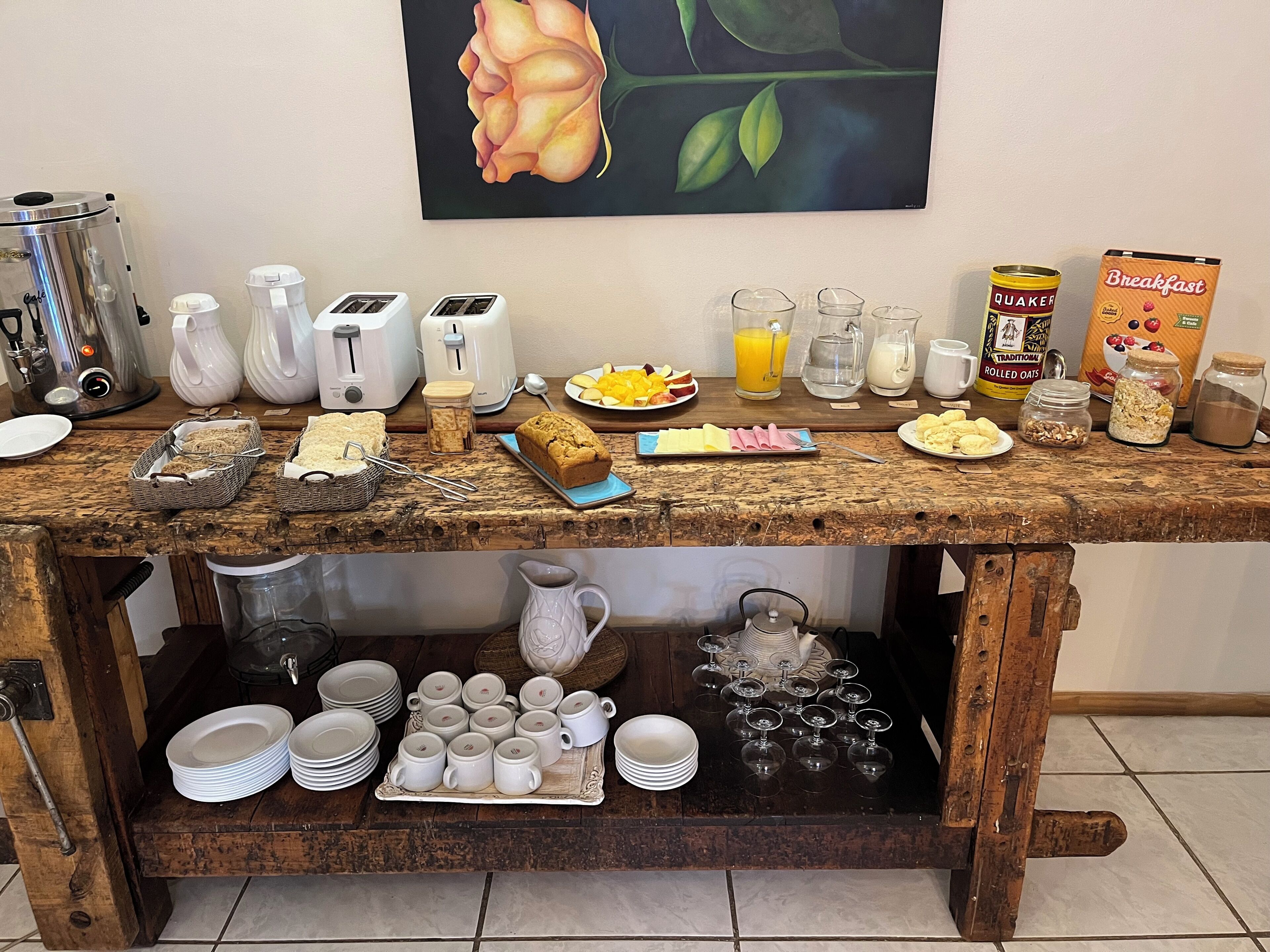 Mesa de desayuno buffet con diversos alimentos, utensilios y un cuadro en la pared.