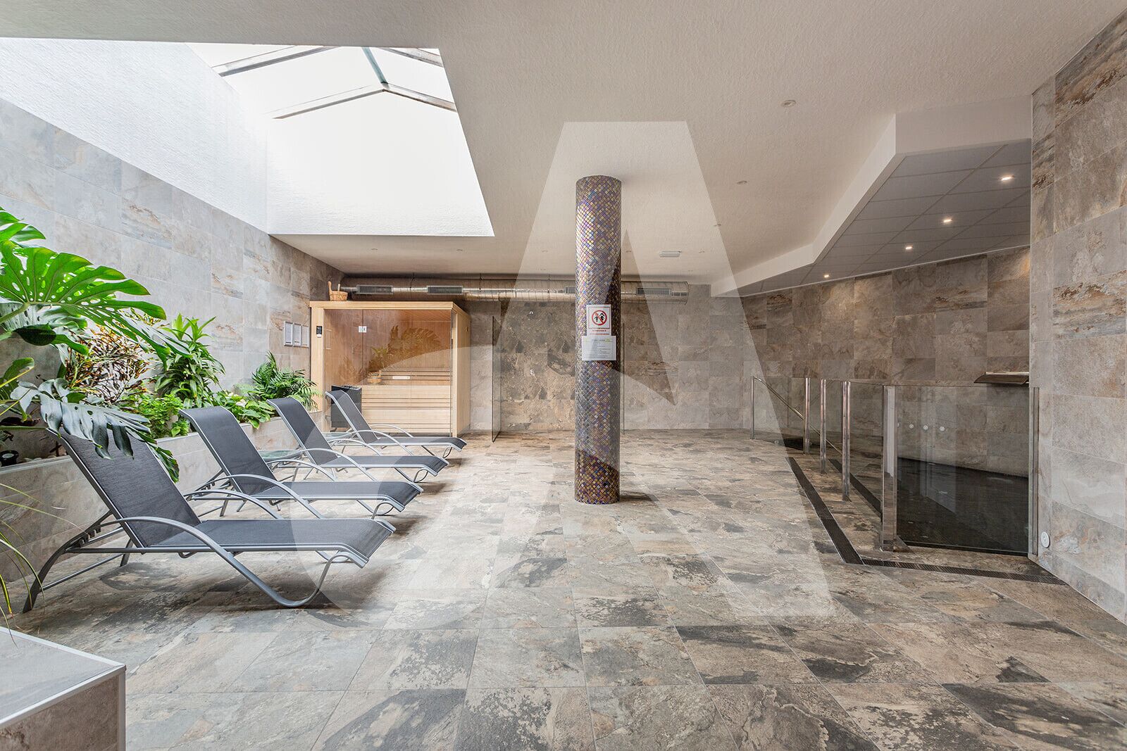 Un área de piscina interior moderna con sillones de descanso, un ascensor de vidrio y un tragaluz.