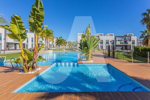 Une piscine dotée d’une terrasse en bois, entourée de bâtiments modernes et de plantes tropicales.