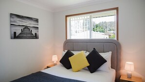 Een hotelkamer met een bed, nachtkastje met lamp, raam met jaloezieën en een schilderij aan de muur.