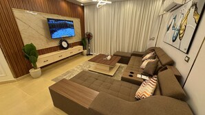 Uma sala de estar moderna com sofá em forma de L, mesa de centro, TV fixada em uma parede de mármore, planta em vaso e uma grande janela com cortinas.