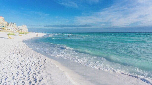 Une plage de sable blanc, une eau turquoise et limpide, et quelques bâtiments visibles au loin.