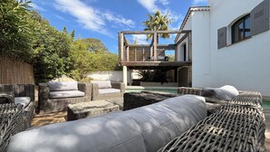 Un patio con mobiliario de mimbre, una piscina y un edificio blanco con contraventanas.