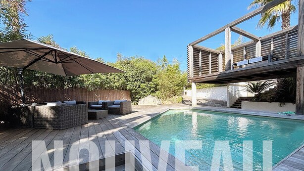Un área junto a la piscina con cubierta de madera, mobiliario exterior y una pérgola con toldo.