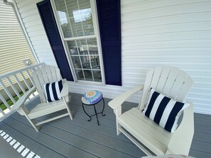 Eine Veranda mit zwei weißen Adirondack-Stühlen, einer mit einem gestreiften Kissen und der andere mit einem einfarbigen Kissen, und einem kleinen runden Tisch mit einer bunten Mosaikplatte.