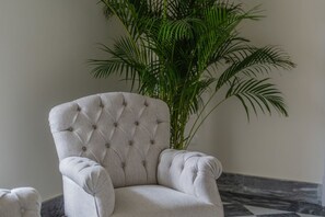 A tufted armchair with a high back and armrests, positioned next to a large potted plant with long, slender leaves. - Signature Villa Overlooking the Gulf of Naples with biolake and garden (Vico Equense)