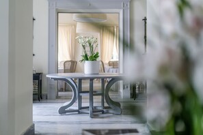 Un área de comedor con una mesa y sillas, una planta en maceta y vista a una habitación con una cama y cortinas.