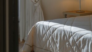 A neatly made bed with a quilted cover, a bedside table with a lamp, and a window with curtains. - Signature Villa Overlooking the Gulf of Naples with biolake and garden (Vico Equense)