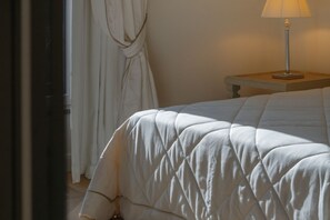 A neatly made bed with a quilted cover, a bedside table with a lamp, and a window with curtains. - Signature Villa Overlooking the Gulf of Naples with biolake and garden (Vico Equense)