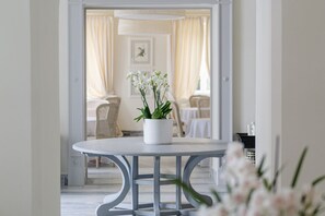 Une table blanche avec une plante en pot au premier plan, un coin repas avec des chaises et une table à l’arrière-plan, et un tableau encadré au mur.