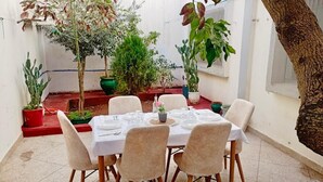 Een eetruimte met een ronde tafel gedekt voor zes personen, omringd door zes stoelen. Achtergrond met potplanten en een boom.