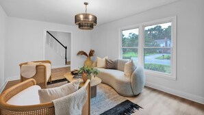A living room with a curved grey sofa, two wicker chairs, a coffee table, and a large window with a view of a house and greenery. - Casa Azul an interior designed home in Stonehaven.Security System,Fiber Internet (Charlotte)