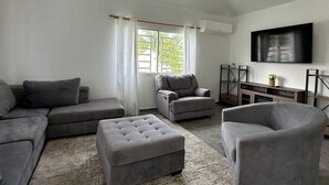 Una sala de estar moderna con sofás grises, un sillón gris y un reposapiés a juego. Un televisor de pantalla plana está montado en la pared, y hay una ventana con cortinas.