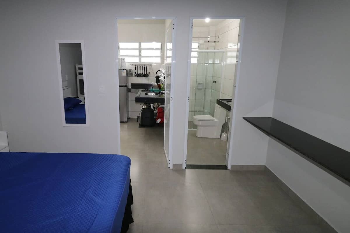Une chambre avec un lit, une petite table de chevet, un miroir et une vue sur la salle de bain et la cuisine.