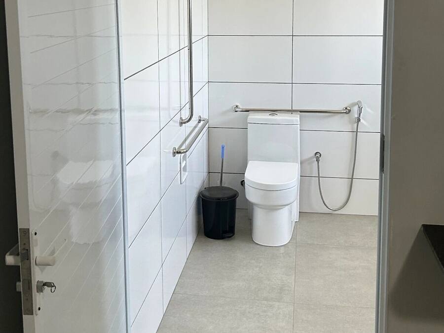 Un baño moderno con azulejos blancos, un inodoro, un cubo de basura y una barra de apoyo.