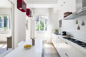 Une cuisine moderne avec des meubles blancs, une grande fenêtre et un coin repas comprenant une table blanche et un saladier rempli de citrons.
