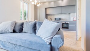 A modern living room with a light blue sofa, a kitchen with grey cabinets, and a large window providing natural light. - Easylife - Style and balcony near the Duomo (Milano)