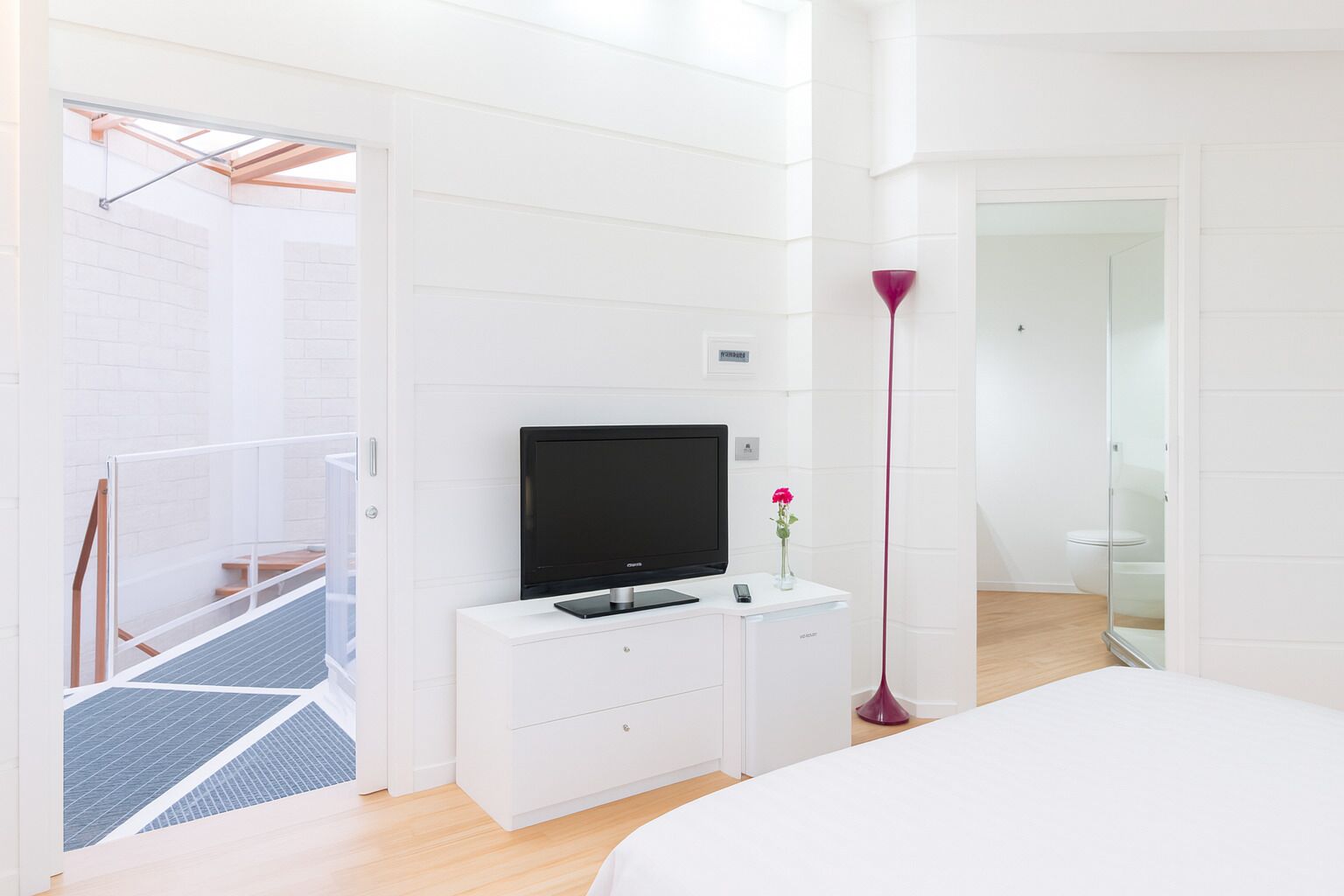 Ein modernes Schlafzimmer mit einem Bett, einem Fernseher auf einer weißen Kommode, einer roten Stehlampe und Blick ins Badezimmer.