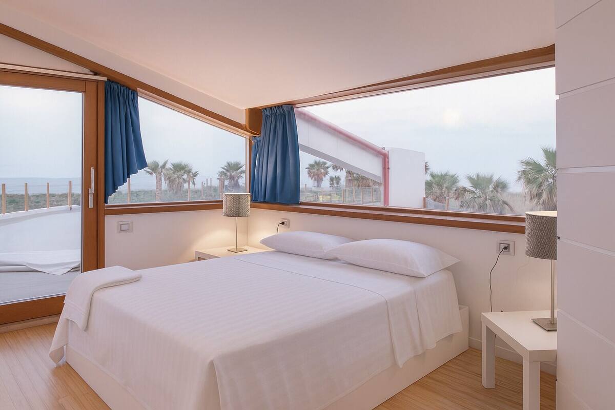 A modern bedroom with a large bed, a bedside table, a lamp, and a view of palm trees through large windows.