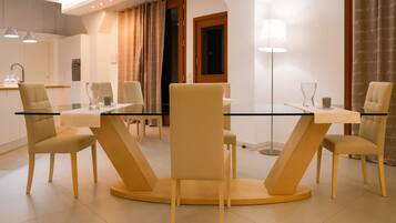 A modern dining area with a glass table and beige chairs, a kitchen in the background, and a floor lamp.