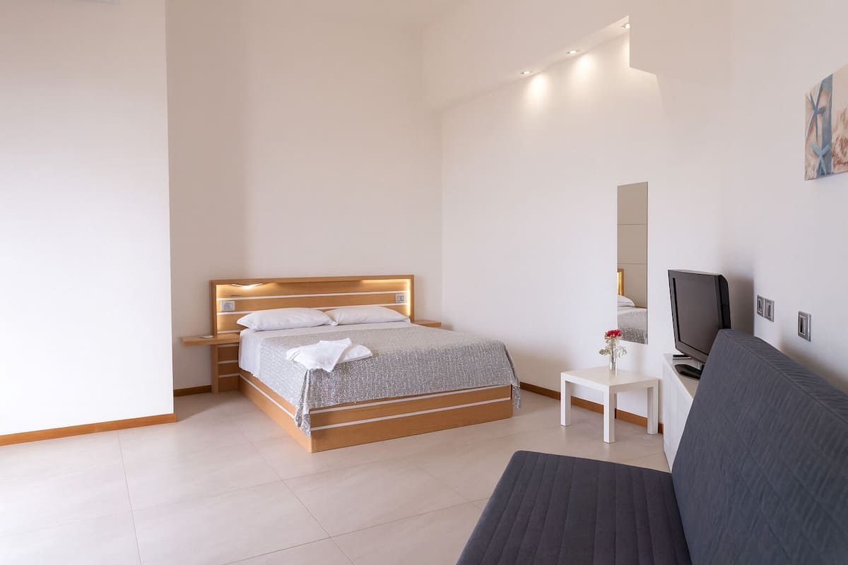 A modern bedroom with a wooden bed, bedside tables, a small white table with a TV, and a grey sofa.