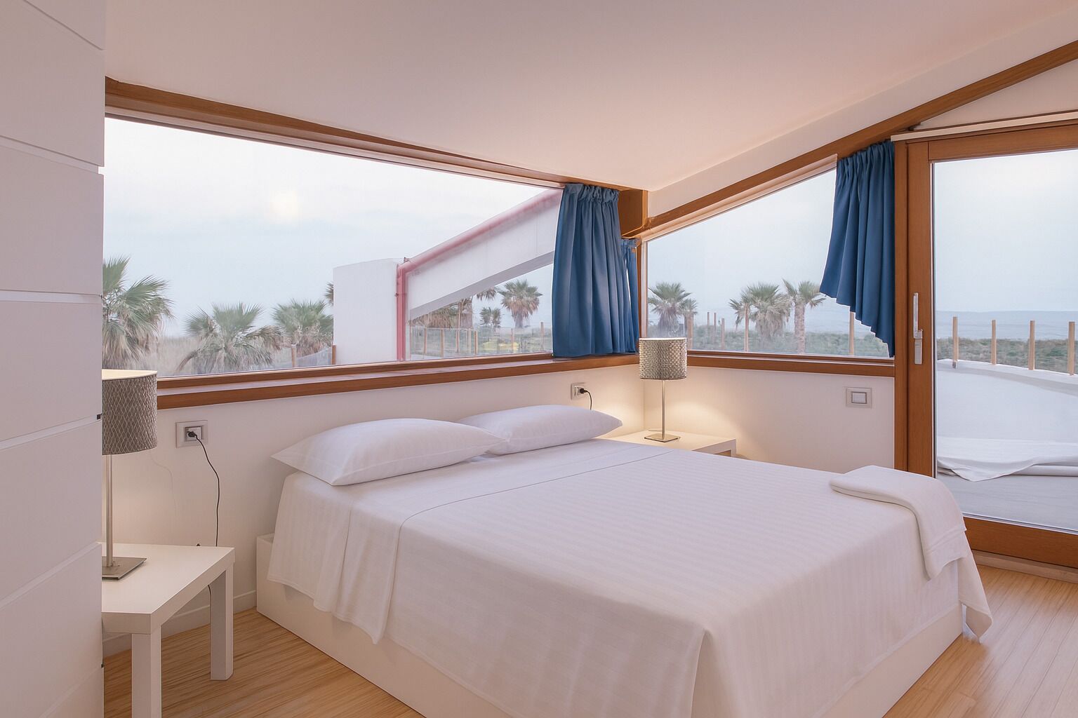 A modern bedroom with a large bed, two bedside tables, a lamp, and a view of palm trees through large windows.