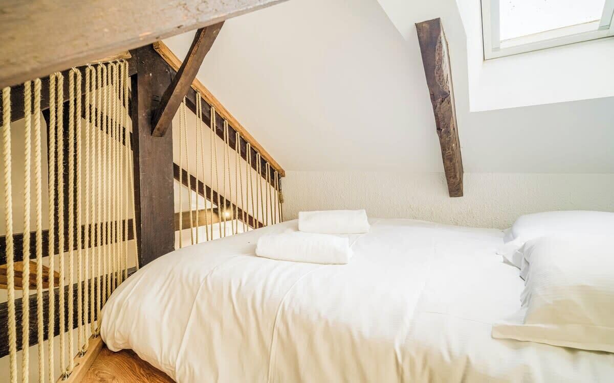A bedroom with a wooden staircase, a bed with white bedding, and a window with a view of the outside.