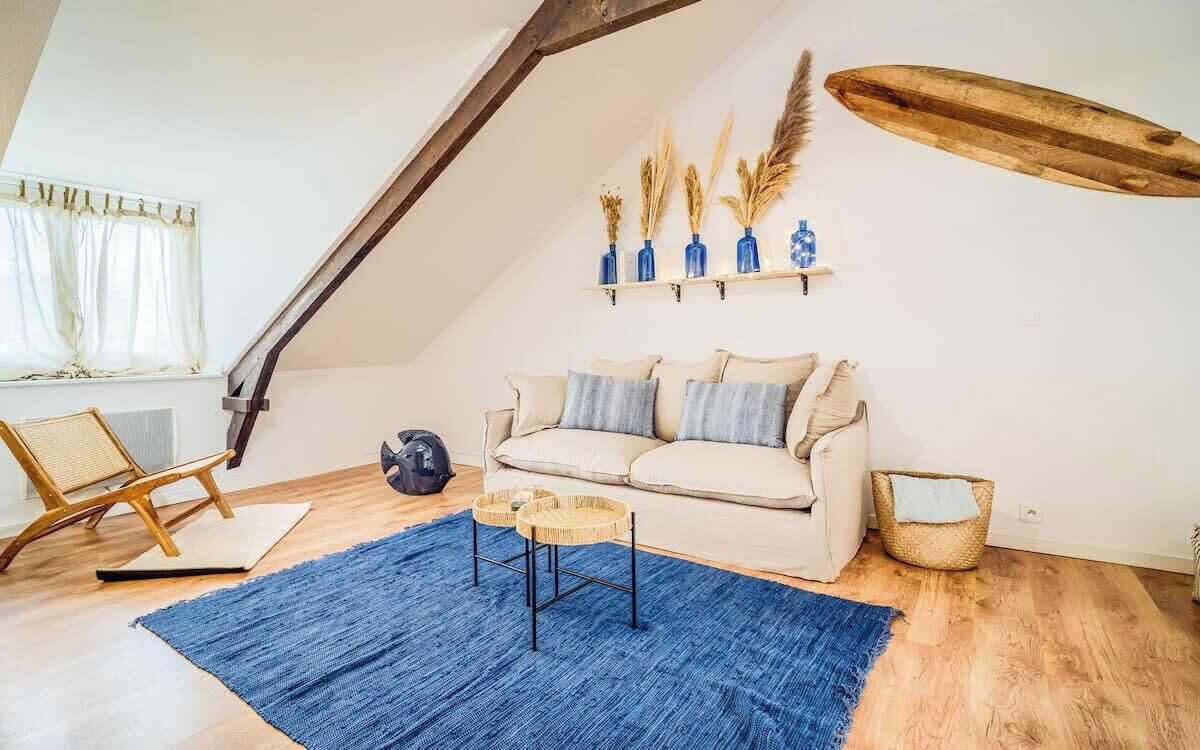 Et loftrum med skrå væg, beige sofa, blå måtte og synlig træbjælke i loftet.