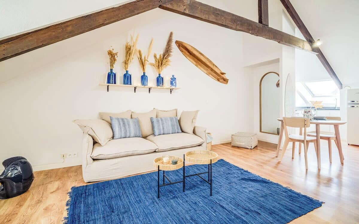 A cozy loft-style room with a high ceiling, exposed wooden beams, a sofa with cushions, a blue rug, and a small table with a basket.